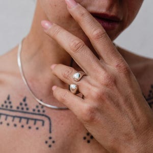 May include: A silver ring featuring two rounded, teardrop-shaped elements. The ring is worn on a finger, and a silver necklace and small hoop earrings are also visible. The person is wearing a light green spaghetti strap top.