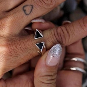 May include: A silver ring with two black triangle-shaped stones. The ring is worn on a finger, and the image shows a close-up view of the jewelry. The ring's design is modern and geometric, with a minimalist aesthetic.