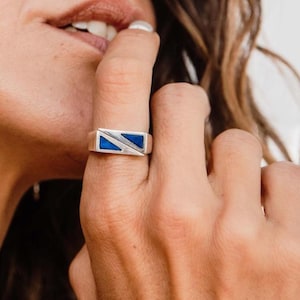 May include: A silver band ring featuring a flat rectangular face with two triangular blue stone inlays separated by a diagonal silver line. The ring has a polished metallic finish and a geometric design that sits horizontally across the finger.