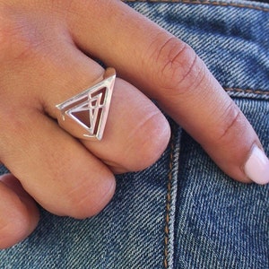 May include: A silver ring with a geometric triangle design. The ring features a hollowed-out triangle shape with an inner geometric pattern. The ring is worn on a finger, with a denim background.