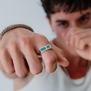 Könnte beinhalten: Ein silberner Ring mit zwei rechteckigen Einlagen aus leuchtend türkisfarbenem Stein. Der Ring hat ein flaches, breites Banddesign und wird an einem Finger getragen. Ein geflochtenes Silberkettenarmband ist ebenfalls am Handgelenk sichtbar.