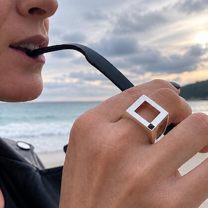 Op de afbeelding: Een zilveren ring met een vierkant geometrisch ontwerp en een klein zwart accent. De ring wordt aan een vinger gedragen, met een zwart voorwerp in de mond. De achtergrond toont een strand en de oceaan.