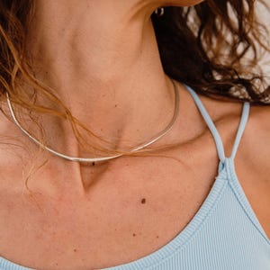 Może przedstawiać: Srebrny naszyjnik typu choker na szyi osoby. Naszyjnik to cienka, prosta opaska. Osoba ma na sobie jasnoniebieski prążkowany top na ramiączkach. Tło jest w neutralnym kolorze.