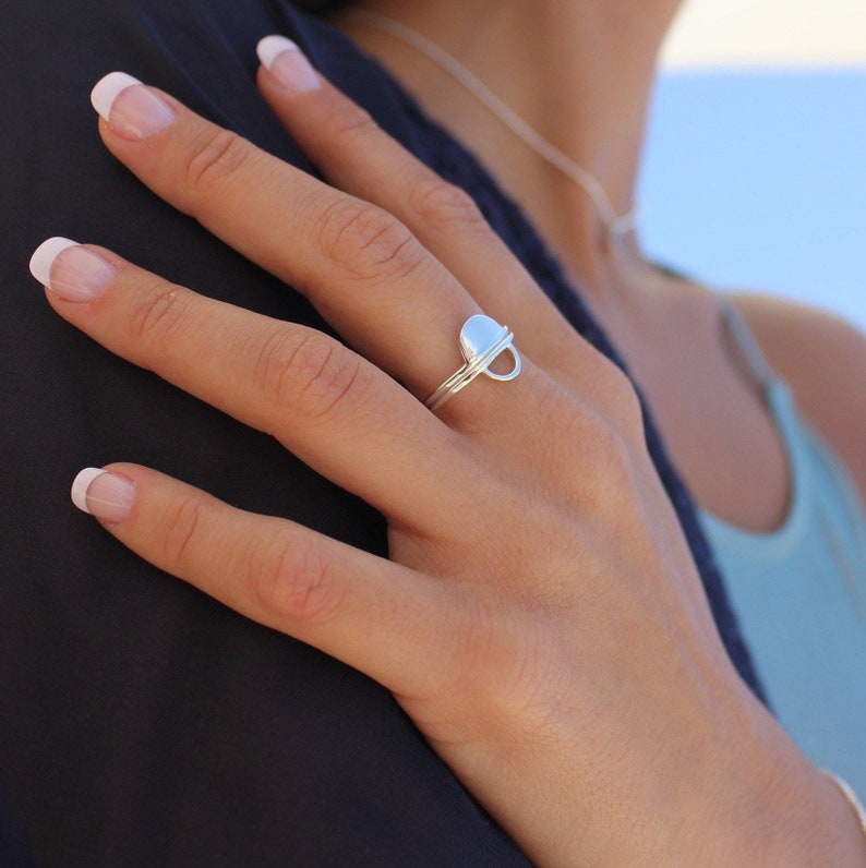 May include: A silver ring with a unique, rounded design featuring a small circle cutout. The ring is worn on the middle finger of a hand.