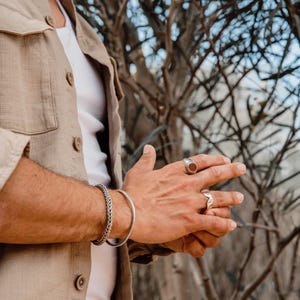K&ouml;nnte beinhalten: Ein silbernes geflochtenes Kettenarmband und ein glatter silberner Armreif am Handgelenk, kombiniert mit zwei Silberringen an den Fingern, einer mit einem runden rotbraunen Stein und der andere mit einem strukturierten, kantigen Design.