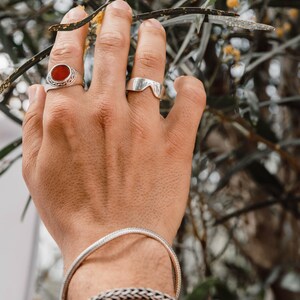 K&ouml;nnte beinhalten: Ein Silberring mit einem runden orangefarbenen Stein, ein schlichter Silberring mit V-f&ouml;rmigem Ausschnitt, ein d&uuml;nnes Schlangenkettchen aus Silber und ein dickeres geflochtenes Silberarmband. Der Schmuck wird an einer Hand getragen.