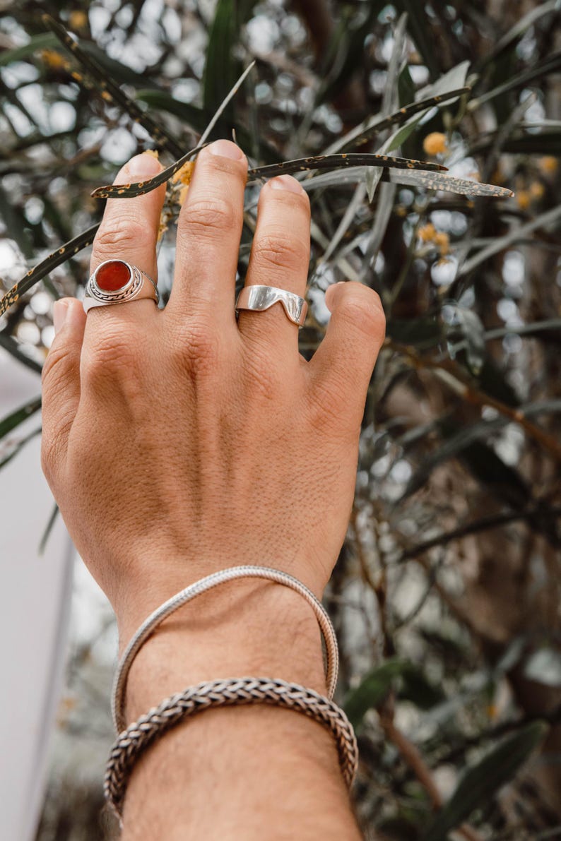 K&ouml;nnte beinhalten: Ein Silberring mit einem runden orangefarbenen Stein, ein schlichter Silberring mit V-f&ouml;rmigem Ausschnitt, ein d&uuml;nnes Schlangenkettchen aus Silber und ein dickeres geflochtenes Silberarmband. Der Schmuck wird an einer Hand getragen.
