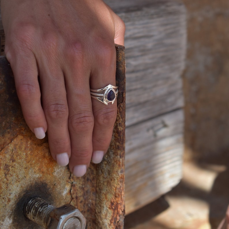 May include: A silver ring with a dark purple gemstone set in a triangular shape. The ring is worn on a finger.