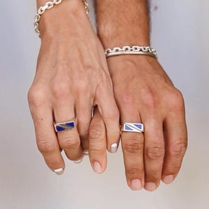 Può includere: Due mani con bracciali a catena d'argento e anelli abbinati. Gli anelli hanno un design rettangolare con un intarsio blu. Le mani si tengono, mettendo in mostra i gioielli.