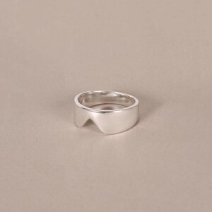 K&ouml;nnte beinhalten: Ein Ring aus poliertem Silber mit einer glatten, breiten Ringschiene, die am oberen Rand eine markante V-f&ouml;rmige Aussparung aufweist. Das Metall hat eine reflektierende Hochglanzoberfl&auml;che und das Design beh&auml;lt eine gleichm&auml;&szlig;ige St&auml;rke bei.