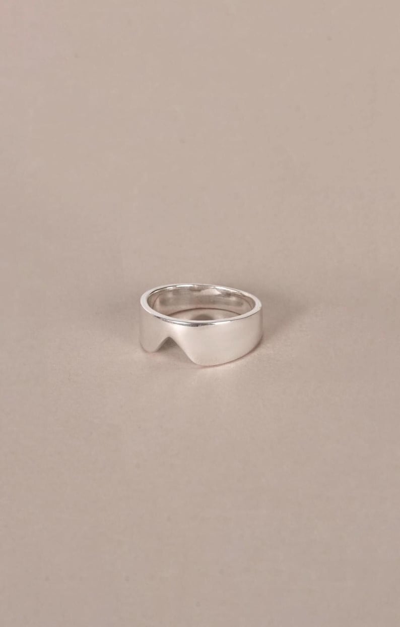 K&ouml;nnte beinhalten: Ein Ring aus poliertem Silber mit einer glatten, breiten Ringschiene, die am oberen Rand eine markante V-f&ouml;rmige Aussparung aufweist. Das Metall hat eine reflektierende Hochglanzoberfl&auml;che und das Design beh&auml;lt eine gleichm&auml;&szlig;ige St&auml;rke bei.