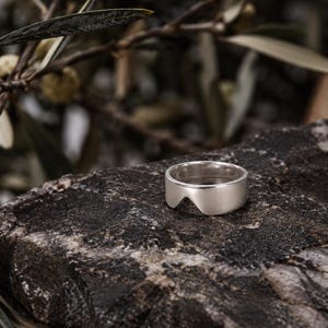 K&ouml;nnte beinhalten: Ein silberfarbener Ring mit glatter, polierter Oberfl&auml;che und einem markanten, bogenf&ouml;rmigen Ausschnitt an der Unterkante der Ringschiene. Der Ring zeichnet sich durch eine breite, massive Form mit einer klaren Oberfl&auml;che aus.