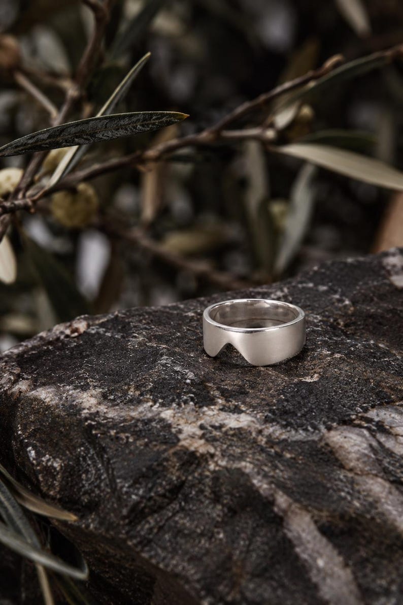 K&ouml;nnte beinhalten: Ein silberfarbener Ring mit glatter, polierter Oberfl&auml;che und einem markanten, bogenf&ouml;rmigen Ausschnitt an der Unterkante der Ringschiene. Der Ring zeichnet sich durch eine breite, massive Form mit einer klaren Oberfl&auml;che aus.