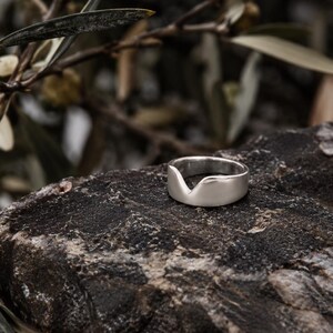 K&ouml;nnte beinhalten: Ein silberfarbener Ring mit glatter, polierter Oberfl&auml;che und einer markanten V-f&ouml;rmigen Einkerbung am oberen Rand der Ringschiene. Der Ring hat eine breite, massive Form mit einer geschwungenen Silhouette, die in der Mitte abf&auml;llt.