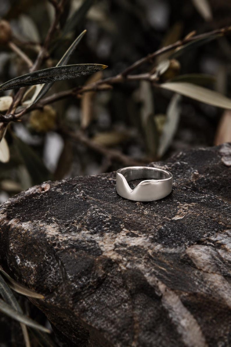 K&ouml;nnte beinhalten: Ein silberfarbener Ring mit glatter, polierter Oberfl&auml;che und einer markanten V-f&ouml;rmigen Einkerbung am oberen Rand der Ringschiene. Der Ring hat eine breite, massive Form mit einer geschwungenen Silhouette, die in der Mitte abf&auml;llt.