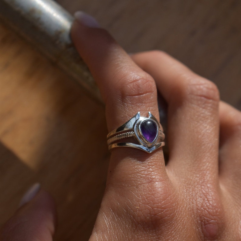 May include: A silver ring featuring a teardrop-shaped amethyst gemstone set in a delicate, pointed design. The ring is worn on a finger.