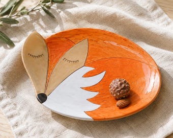 May include: An oval ceramic dish shaped like a sleeping fox with orange, white, and tan colors. The fox face features closed eyes with eyelashes and a black nose. The surface has small dash-like markings throughout the orange area of the dish.