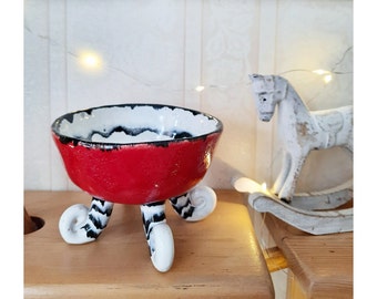 Puede incluir: Un cuenco de cerámica hecho a mano con un exterior rojo vibrante y un interior blanco con detalles en negro. El cuenco está sostenido por tres patas blancas con detalles a rayas blancas y negras. Un caballo balancín decorativo está en el fondo.
