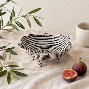 May include: A shallow ceramic bowl with a scalloped rim and three short legs, featuring a hand-painted pattern of thin, concentric black lines on a white background. The bowl has a textured, organic shape and is finished with a glossy glaze.