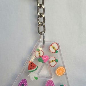 May include: A clear resin keychain shaped like the letter A with a silver chain. The keychain is filled with colorful fruit and mushroom charms.