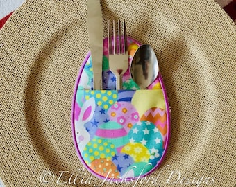 May include: A colourful Easter egg-shaped fabric holder with a bunny design, holding a fork, knife, and spoon. The holder is sitting on a brown burlap placemat.