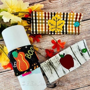 May include: Three different fabric covers for a tumbler or water bottle. The first cover is plaid with a yellow leaf applique. The second cover is black with a pumpkin and the word "Boo" on it. The third cover is white with a red apple and a wood grain pattern.