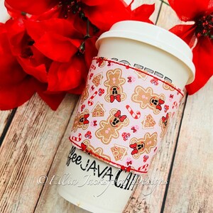 May include: A white coffee cup with a red and white gingerbread man themed sleeve. The sleeve has the text "Coffee Java Cup" on it. The cup is surrounded by red poinsettia flowers.
