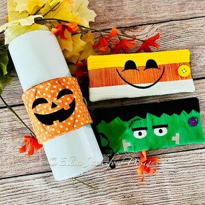 May include: Three Halloween-themed fabric cup cozies. One is orange with a jack-o'-lantern face, one is green with a Frankenstein face, and one is orange and yellow with a smiling face.