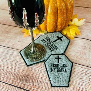 Puede incluir: Dos posavasos bordados en gris y negro con forma de ataúd, con una cruz y el texto "Here Lies My Drink". Un vaso negro con un tallo de mano de esqueleto plateado está encima de uno de los posavasos. Una pequeña calabaza naranja está en el fondo.