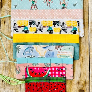 May include: Three fabric pencil rolls with different patterns. The top roll has a light blue background with a pattern of women in dresses and the text "Sew Over It". The middle roll has a white background with a pattern of men in overalls and the text "68 Special". The bottom roll has a pink and white gingham pattern with a watermelon slice on top.