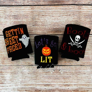 May include: Three black Halloween-themed can coolers. The first cooler says "Gettin' Sheet Faced" with a ghost. The second cooler says "Let's Get Lit" with a jack-o-lantern. The third cooler says "Booty & Booze" with a skull and crossbones.