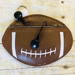 Pode incluir: Uma bolsa de couro marrom em forma de bola de futebol com zíper e costuras brancas, apresentando um design de cadarço de futebol branco. Os fones de ouvido pretos estão guardados dentro da bolsa.