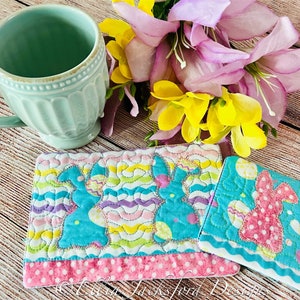 May include: Two quilted fabric coasters featuring a bunny design with Easter eggs. The coasters are in shades of pink, blue, and green. One coaster has a pink bunny and the other has a blue bunny.