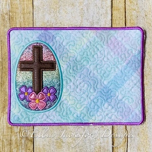 May include: A light blue quilted fabric square with a purple border. The center features a glittery Easter egg with a brown cross and pink and purple flowers.