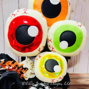 May include: Four plush eyeball pillows in different colors: orange, red, green, and yellow. The pillows are arranged in a pyramid shape on a wooden shelf. The pillows are decorated with black stitching and white accents. The pillows are surrounded by fall decor, including black roses, white pumpkins, and orange leaves.