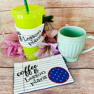 May include: A white coffee cup cozy with blue stitching and the text "coffee & lesson plans" in black. A blue and white polka dot circle is on the right side of the cozy. The cozy is on a wooden surface.