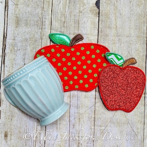 May include: A light blue ceramic teacup next to two apple-shaped appliqués. One apple is red with green polka dots, and the other is red glitter. The appliqués have green leaves and brown stems. The background is a light wood grain.