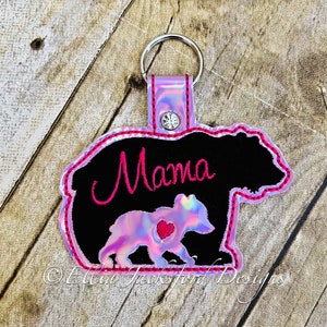 May include: A pink and black bear-shaped keychain with the word "Mama" embroidered in pink on the bear's back. The bear has a pink heart on its chest.