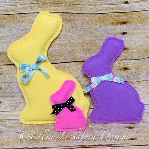 May include: Three felt Easter bunny shapes. The largest bunny is yellow with a blue and white ribbon. The middle bunny is pink with a black and white polka dot ribbon. The smallest bunny is purple with a blue, white, and pink ribbon.