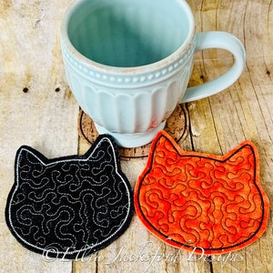 Puede incluir: Dos posavasos con forma de gato, uno negro y otro naranja, con un patrón de espiral. Los posavasos están sobre una superficie de madera, con una taza azul claro encima de uno de ellos.
