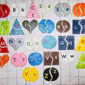 May include: A set of 26 alphabet puzzle pieces, each with a different letter of the alphabet. The pieces are made of felt and are in various colors, including red, green, blue, yellow, orange, pink, and brown. Each piece is cut in half, with the letter on one half and the lowercase letter on the other half. The pieces are arranged in rows on a white wooden surface.