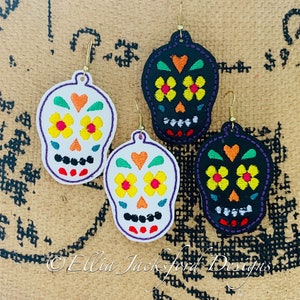 May include: A pair of sugar skull earrings with colorful embroidery. The earrings are made of white and black leather and have gold hooks. The embroidery features yellow flowers, orange hearts, and green leaves.