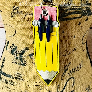May include: A yellow pencil-shaped badge holder with a pink eraser top and two blue pens inside. The badge holder has a silver ring at the top for attaching a lanyard.