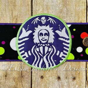 May include: A purple and white coffee cup cozy with a green border. The design features a white silhouette of a character with a skull and crossbones on the top. The text "Ellia Jackford Designs" is visible at the bottom of the image.