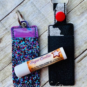 Puede incluir: Dos llaveros con un soporte para bálsamo labial. Un llavero es negro con un diseño de vaca y el otro es morado con purpurina y una ventana de plástico transparente. Ambos llaveros tienen un anillo plateado para sujetar las llaves. El bálsamo labial es blanco con una etiqueta amarilla que dice "Sugar Cookie" y tiene un diseño de estrella.