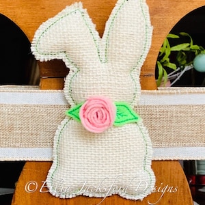 May include: A white burlap bunny with green stitching and a pink felt flower with green leaves. The bunny is sitting on a brown wooden surface with a burlap ribbon.
