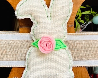 May include: A white burlap bunny with green stitching and a pink felt flower with green leaves. The bunny is sitting on a brown wooden surface with a burlap ribbon.