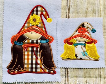 May include: Two embroidered gnome ornaments with fall colors and patterns. The larger gnome has a plaid dress and brown hair. The smaller gnome has a white dress with a floral pattern and yellow hair. Both gnomes have a red hat with a yellow flower.