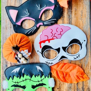 Puede incluir: Cuatro máscaras de fieltro con temática de Halloween: un gato negro, un Frankenstein verde, una calavera blanca con un cerebro rosa y una calabaza. Las máscaras están sobre una superficie de madera con hojas naranjas.