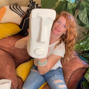 May include: A white ceramic sculpture of a face with a large nose and lips. The sculpture is being held by a person wearing a white shirt and blue jeans.
